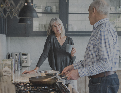 A woman talking to a man holding a cup in a kitchen while some cooking is taking place.